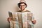 Young blond explorer man with beard and blue eyes wearing glasses and hat holding city map with a happy face standing and smiling