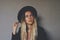 Young blond female wiccan witch wearing a black witch hat and black blazer. Artsy portrait against dark grey wall