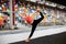 Young blond fit woman, wearing orange top, black leggings, orange sneakers, doing stretching exercise by railing on stadium.