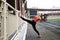 Young blond fit woman, wearing orange top, black leggings, orange sneakers, doing stretching exercise by railing on stadium.