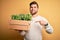 Young blond gardener man with beard and blue eyes holding wooden box with plants very happy pointing with hand and finger