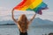 Young blond girl holding  LGBTQI flag above head walking in the day on the white beach near the sea