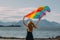 Young blond girl holding  LGBTQI flag above head walking in the day on the white beach near the sea