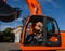 A young blond hair woman sits in a cabin of a new orange crawler excavator with a big black bucket.