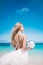 Young blond long hair bride wear a white open back wedding dress and stand on the white sand beach with a pearl. Looking to the se