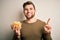 Young blond man with beard and blue eyes holding bowl with unhealthy potatoes chips surprised with an idea or question pointing