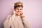 Young blond man with curly hair wearing golden crown of king over pink background covering ears with fingers with annoyed