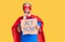 Young blond man wearing super hero costume holding act now cardboard banner serious face thinking about question with hand on