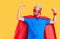 Young blond man wearing super hero custome showing arms muscles smiling proud