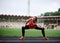 Young blond sportswoman, wearing orange top and black leggings, doing squats on a stadium with green grass in summer. Full-length