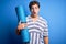 Young blond sporty man with curly hair holding mat to do yoga over isolated blue background scared in shock with a surprise face,