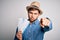 Young blond tourist man with blue eyes on vacation wearing hat holding boarding pass pointing with finger to the camera and to