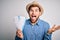 Young blond tourist man with blue eyes on vacation wearing hat holding boarding pass very happy and excited, winner expression