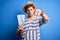 Young blond tourist man with curly hair on vacation wearing summer hat holding boarding pass with angry face, negative sign