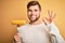 Young blond vegetarian man with beard and blue eyes holding fork with fresh cob corn doing ok sign with fingers, excellent symbol