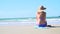 Young blond woman with blue bikini and straw hat sits on the beach and looks at the sea