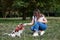 Young blond woman, kneeling down, holding her dog`s paw in park in summer. Dog owner training her Cavalier king charles spaniel