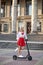 Young blond woman, posing with black electric scooter in city center in front of old historical building with pillars. Summer