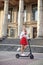 Young blond woman, posing with black electric scooter in city center in front of old historical building with pillars. Summer