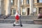 Young blond woman, posing with black electric scooter in city center in front of old historical building with pillars. Summer