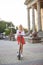 Young blond woman, posing with black electric scooter in city center in front of old historical building with pillars. Summer