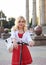 Young blond woman, posing with black electric scooter in city center in front of old historical building with pillars. Summer