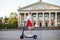 Young blond woman, riding on black electric scooter in city center in front of old historical building with pillars. Summer