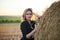 Young blond woman, wearing black jacket and jeans shorts, leaning on wheat bale on rural field on summer evening. Three-quarter