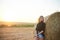 Young blond woman, wearing black jacket and jeans shorts, leaning on wheat bale on rural field on summer evening. Three-quarter