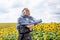 Young blond woman, wearing boho hippie clothes, holding grey transparent shawl scarf, waving on wind on yellow sunflowers field.