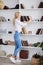 Young blond woman, wearing casual jeans and white top, standing near book shelves in the office room. Freelancer employed at home