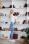 Young blond woman, wearing casual jeans and white top, standing near book shelves in the office room. Freelancer employed at home