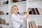 Young blond woman, wearing casual jeans and white top, standing near book shelves in the office room. Freelancer employed at home