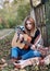 Young blond woman, wearing colorful cardigan and blue jeans, holding acoustic guitar, sitting on blanket in by wooden fence in