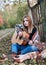 Young blond woman, wearing colorful cardigan and blue jeans, holding acoustic guitar, sitting on blanket in by wooden fence in