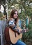 Young blond woman, wearing colorful cardigan and blue jeans, holding acoustic guitar, standing by wooden fence in countryside.