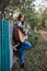 Young blond woman, wearing colorful cardigan and blue jeans, holding acoustic guitar, standing by wooden fence in countryside in