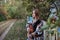 Young blond woman, wearing colorful cardigan and blue jeans, holding acoustic guitar, standing by wooden fence in countryside.