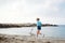 Young blond woman, wearing jeans shorts and turquoise blouse, running at the beach by the sea, holding light grey white husky