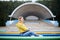 Young blond woman, wearing yellow hoody, blue jeans and eyeglasses, sitting on colorful benche in city urban park in summer.