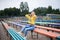 Young blond woman, wearing yellow hoody, blue jeans and eyeglasses, sitting on colorful benche in city urban park in summer.