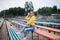 Young blond woman, wearing yellow hoody, blue jeans and eyeglasses, sitting on colorful benche in city urban park in summer.