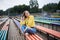 Young blond woman, wearing yellow hoody, blue jeans and eyeglasses, sitting on colorful benche in city urban park in summer.