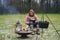 Young blond woman in a wool sweater reads a book outside on the grass by the campfire. Burning fire in fire bowl, a