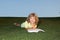 A young blonde boy is lying on the green grass outdoors reading a white book in summer. Smart child reading book, laying