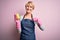 Young blonde cleaner woman with short hair wearing apron and gloves holding scrub scourer with surprise face pointing finger to