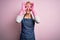 Young blonde cleaner woman with short hair wearing apron and gloves over pink background doing ok gesture like binoculars sticking