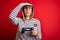 Young blonde gamer woman using gamepad playing video games over red background stressed with hand on head, shocked with shame and