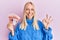 Young blonde girl holding 100 norwegian krone banknotes doing ok sign with fingers, smiling friendly gesturing excellent symbol