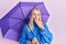 Young blonde girl holding purple umbrella covering mouth with hand, shocked and afraid for mistake